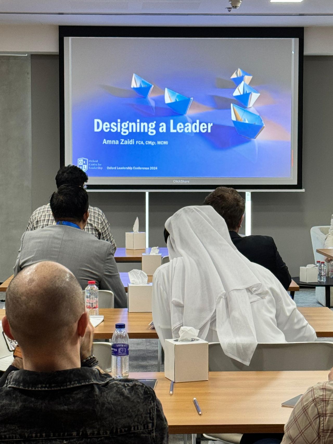 A conference audience listening to a presentation on "Designing a Leader" by Amna Zaidi.